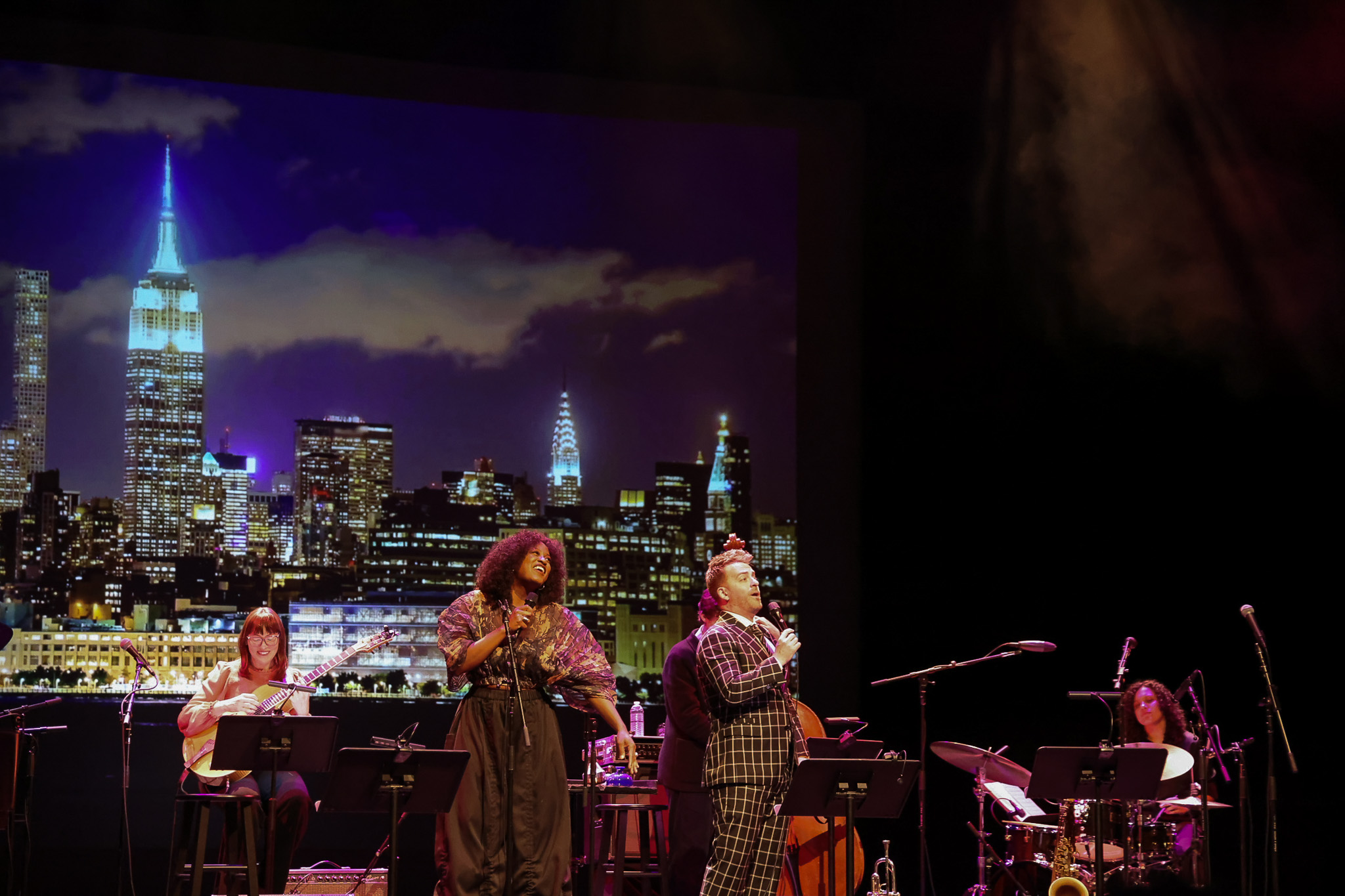 Musicians perform on stage in front of a projected New York City skyline at night, with two vocalists and instrumentalists playing guitar and drums.