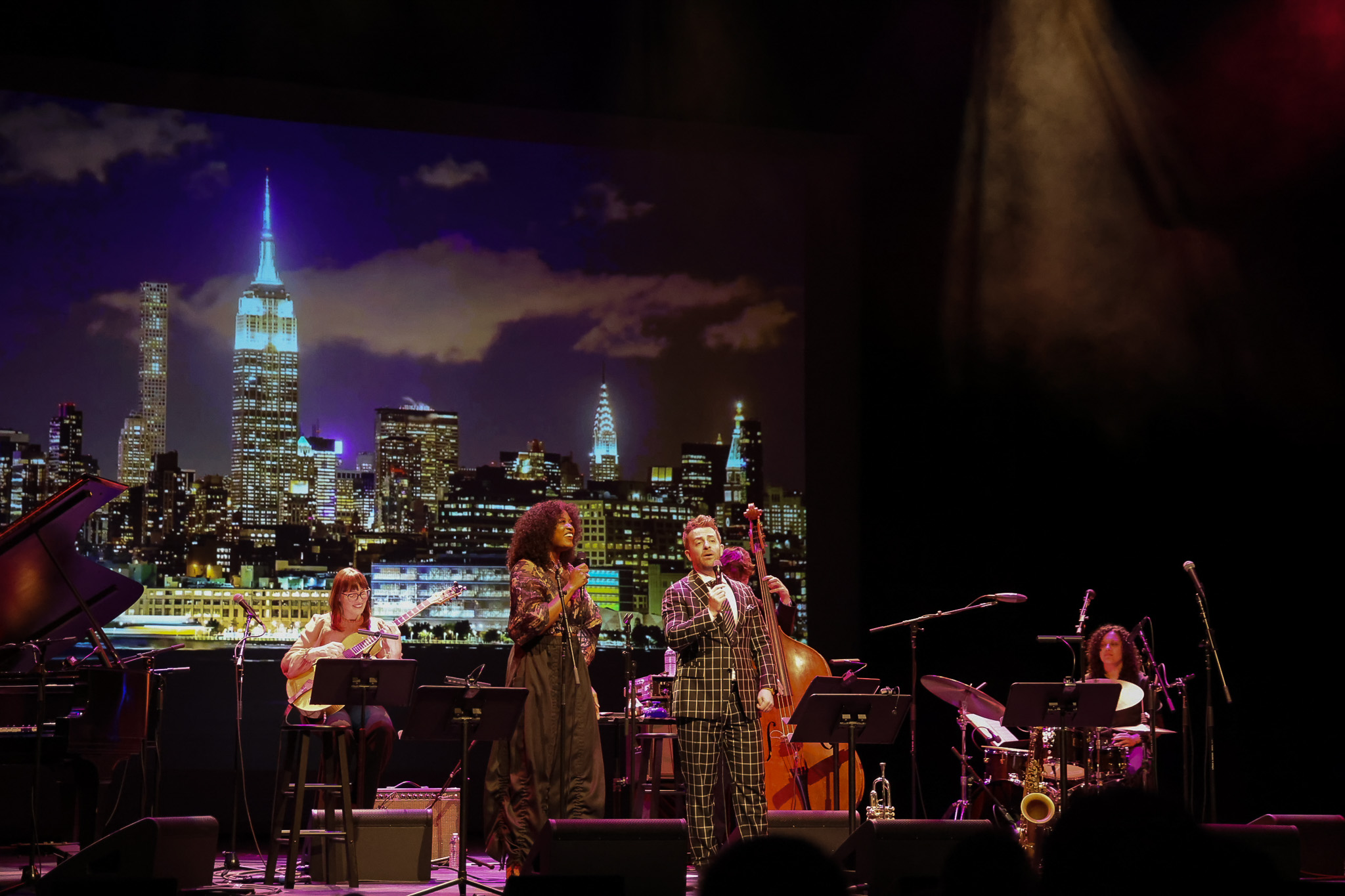 A jazz band performs on stage with a city skyline backdrop, including the Empire State Building lit up at night.