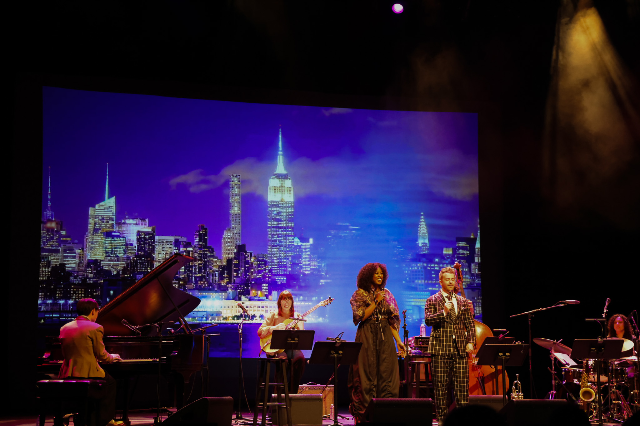 A jazz band performs on stage with a pianist, two vocalists, a bassist, and a drummer; a city skyline is displayed in the background.