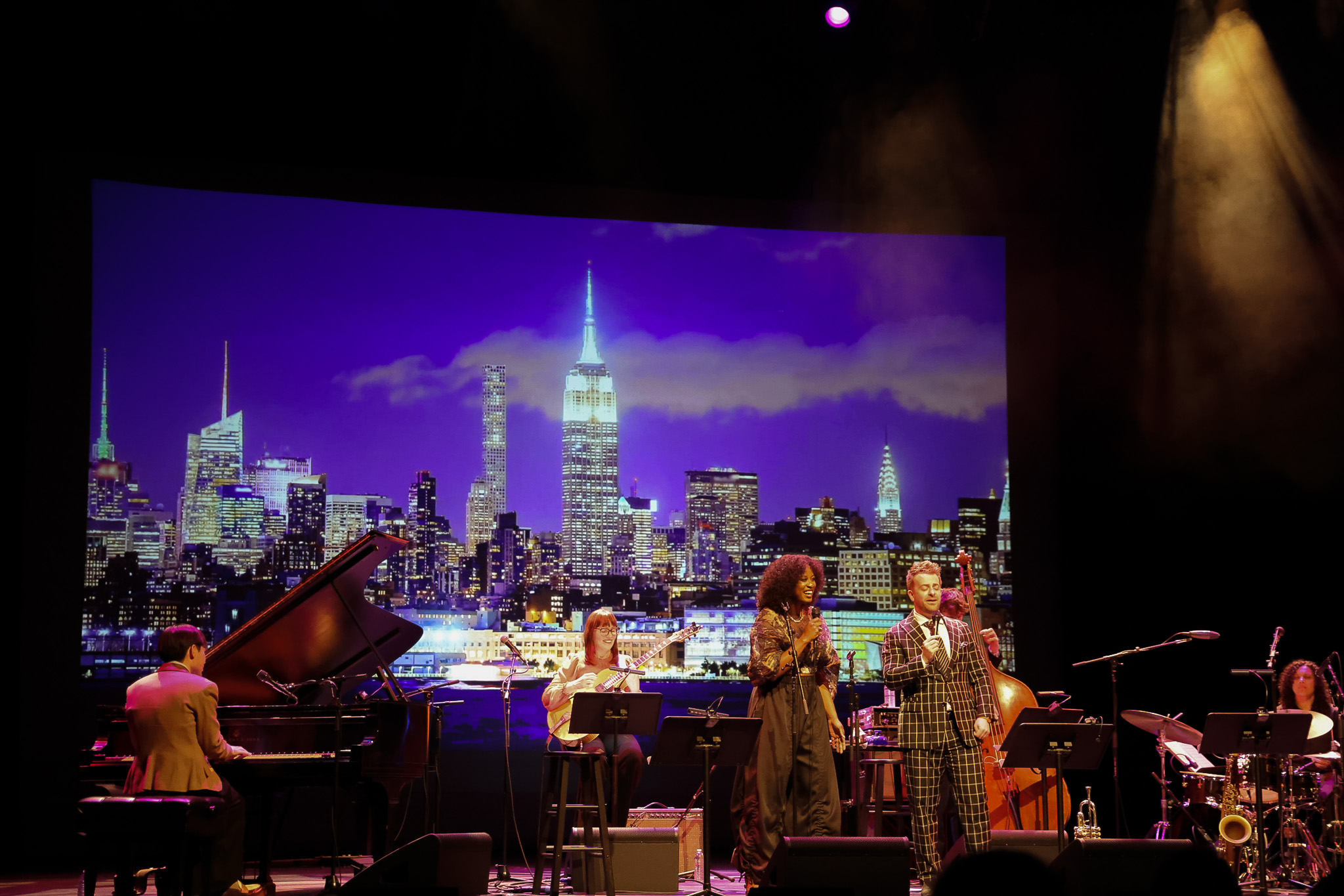 A jazz band performs on stage in front of a large city skyline backdrop featuring the Empire State Building at night.