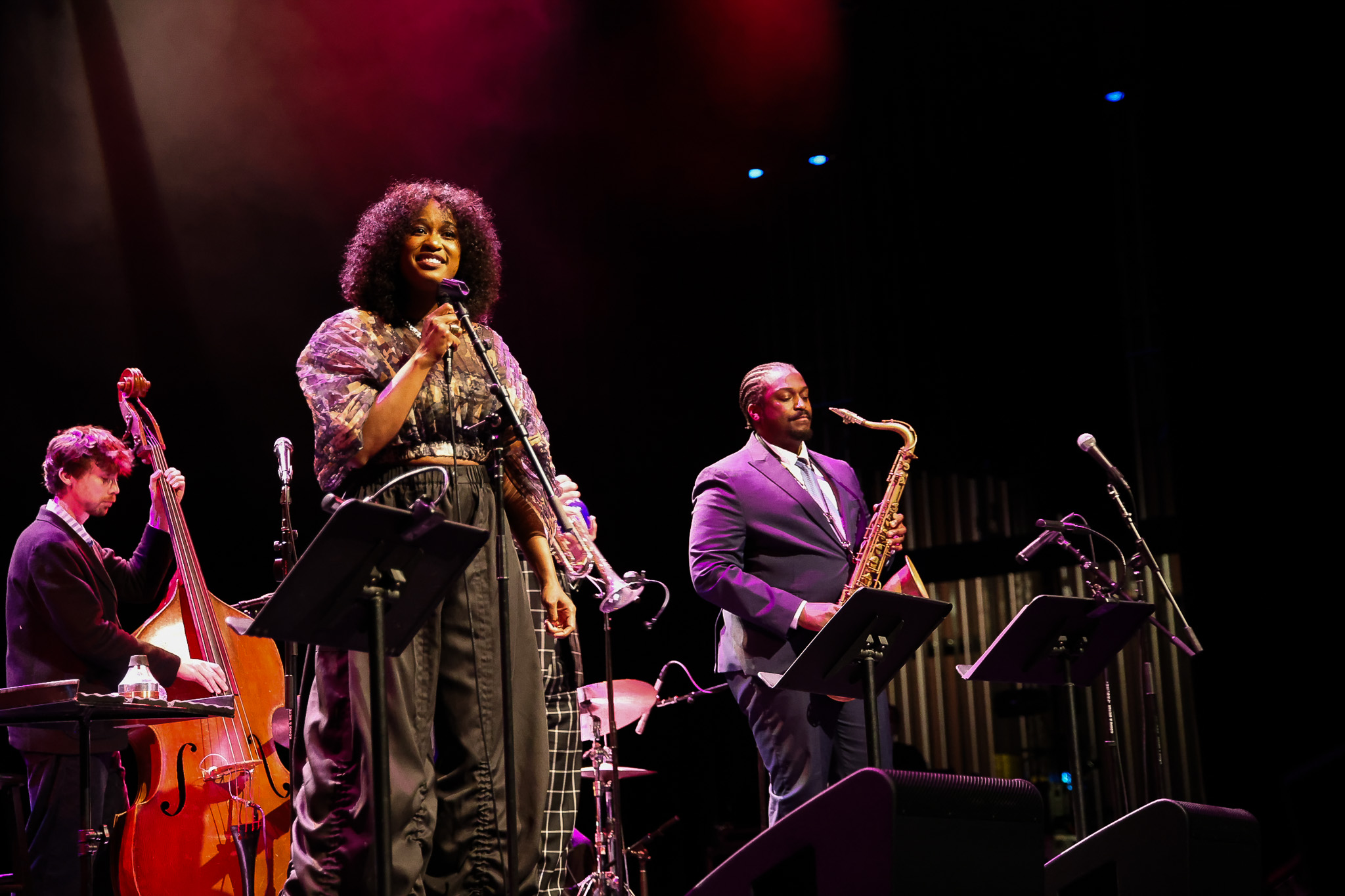 A jazz band performs on stage; a woman sings at the microphone while three male musicians play upright bass, saxophone, and drums under stage lighting.