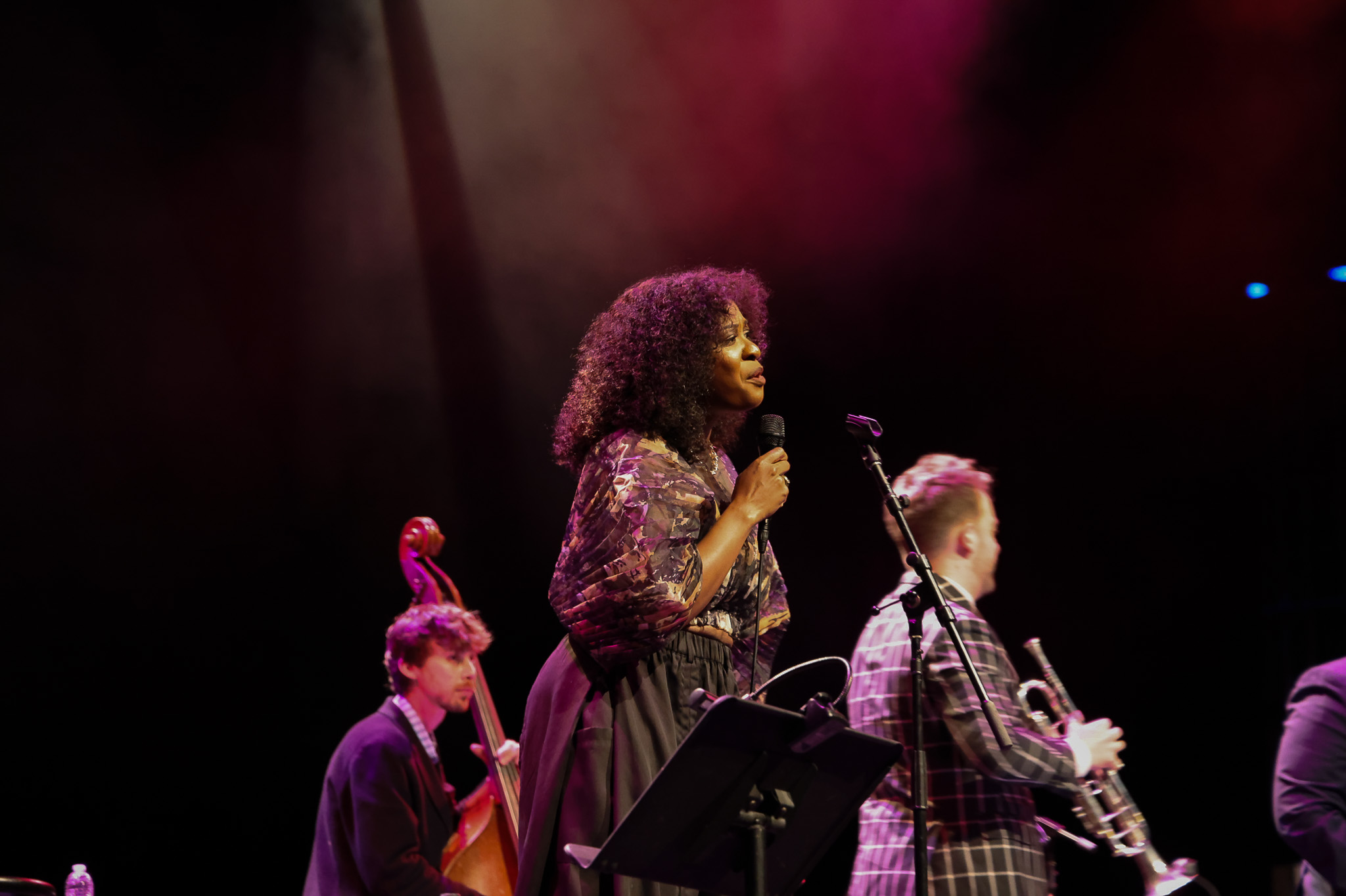 A woman sings into a microphone on stage with a jazz band, including a double bass player and a trumpet player, under colorful stage lights.