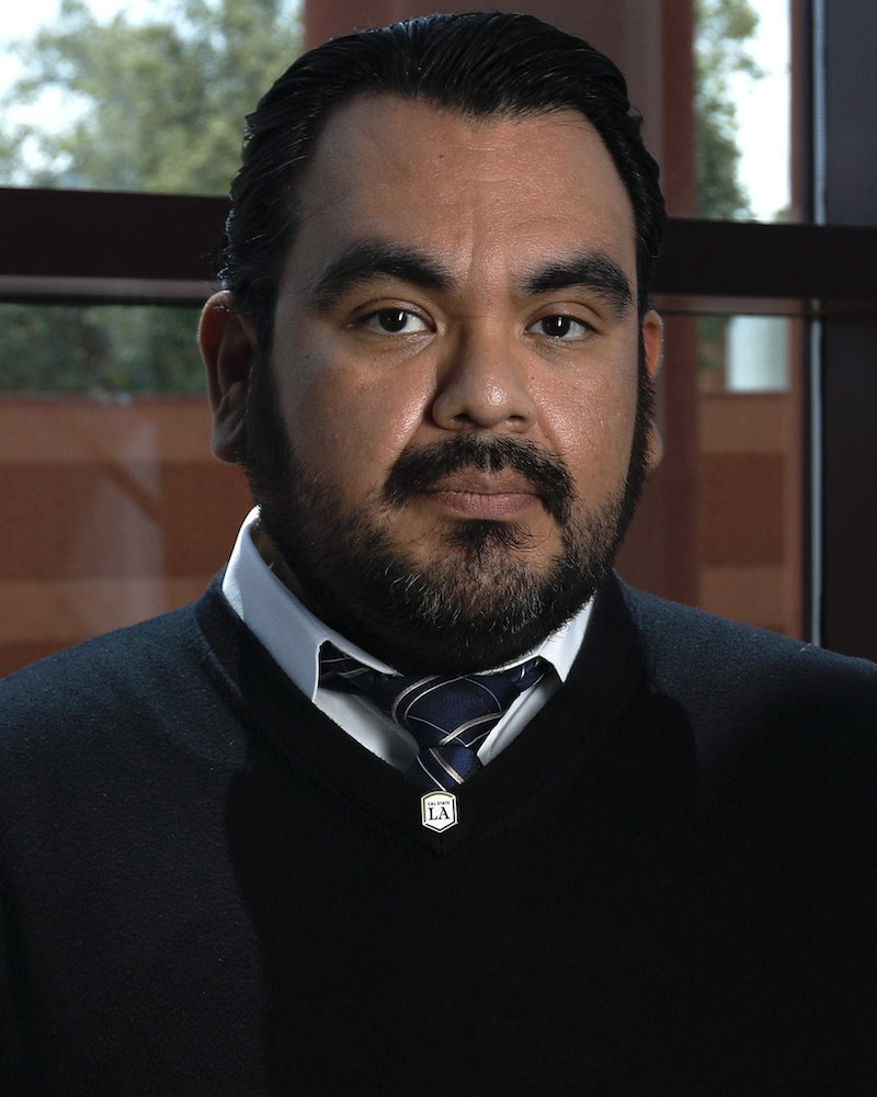 A man with dark hair and a beard wearing a black sweater, white shirt, and tie stands indoors in front of a window.