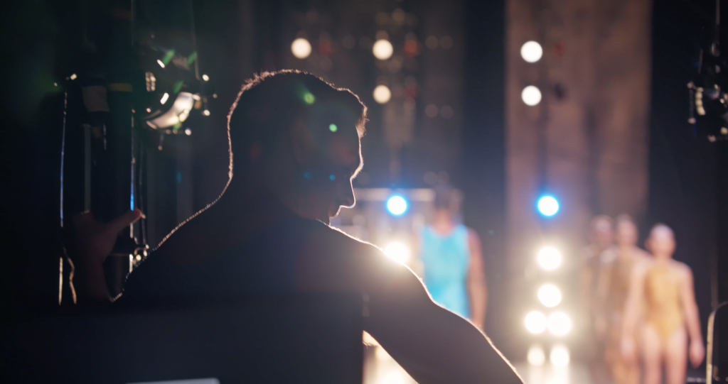 A dancer waits in the wings of a stage to go on stage with lights shining on him