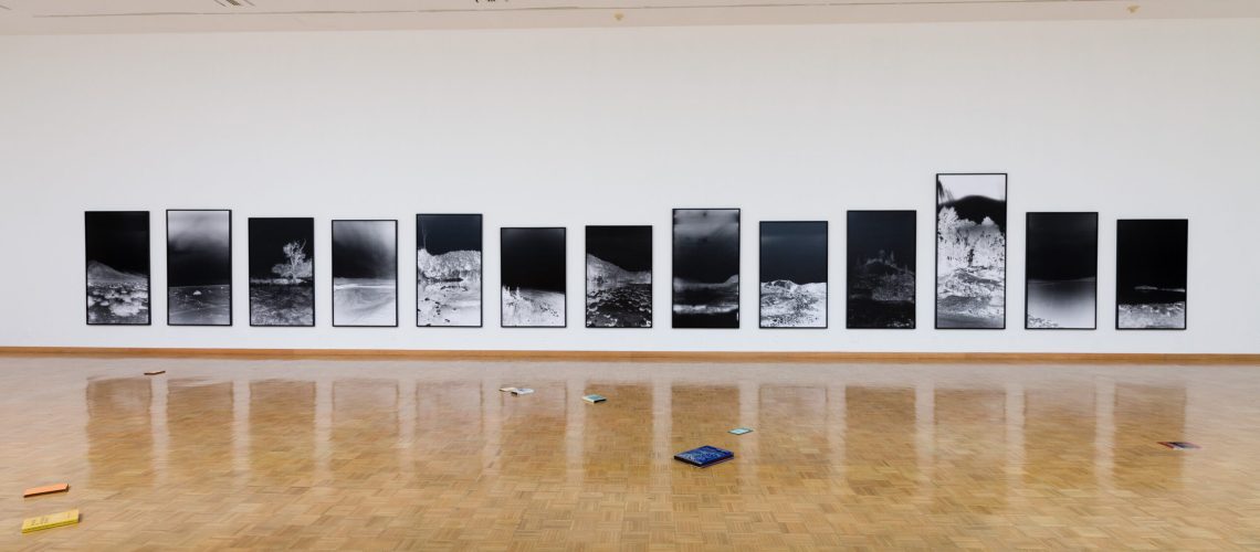 A spacious art gallery with a herringbone wood floor displays a series of large black and white photographic prints in linear arrangement on a clean white wall, with a scattering of colorful objects on the floor in the foreground.