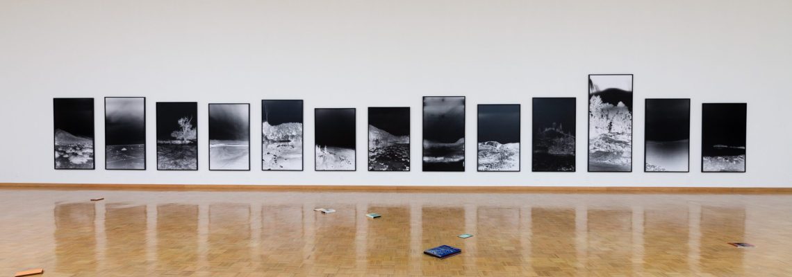 A spacious art gallery with a herringbone wood floor displays a series of large black and white photographic prints in linear arrangement on a clean white wall, with a scattering of colorful objects on the floor in the foreground.