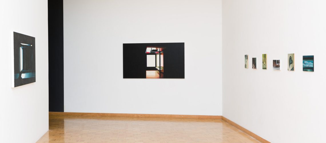 A minimalist art gallery interior with white walls featuring an assortment of framed photographs and paintings displayed at varying heights, with a parquet floor leading up to the artwork.