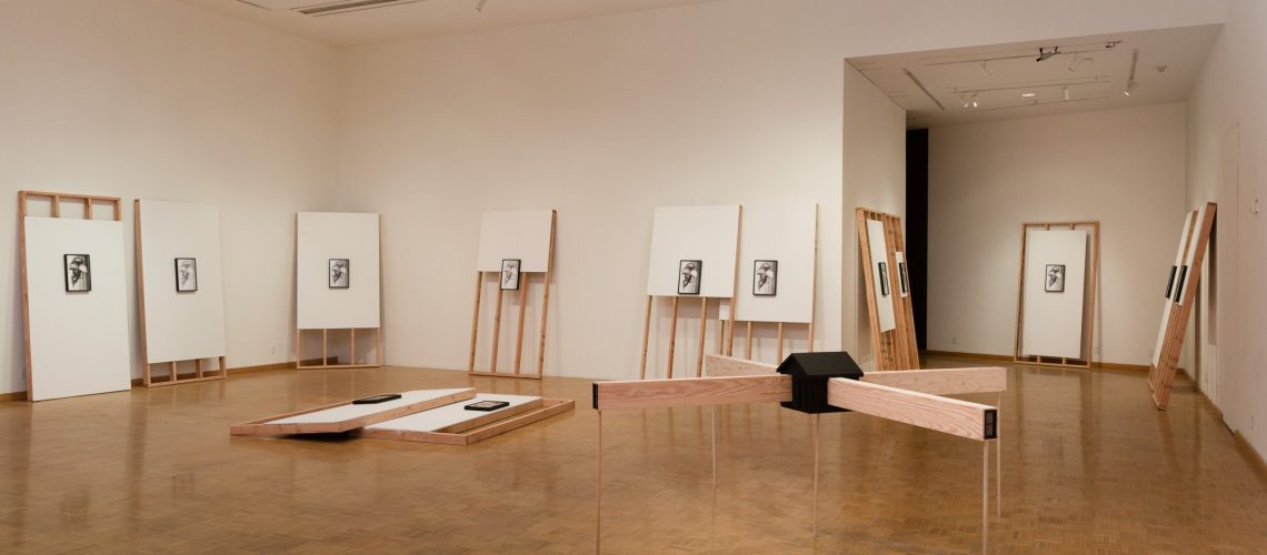 An art gallery room featuring a minimalistic exhibition with several framed artworks displayed on easels and two wooden benches for viewers to sit and contemplate the pieces.