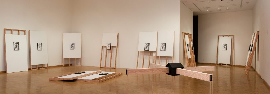 An art gallery room featuring a minimalistic exhibition with several framed artworks displayed on easels and two wooden benches for viewers to sit and contemplate the pieces.