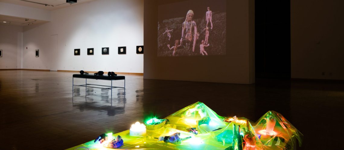 A contemporary art exhibition featuring a video projection and illuminated sculptural installations in a gallery setting, with framed artworks on the walls in the background.