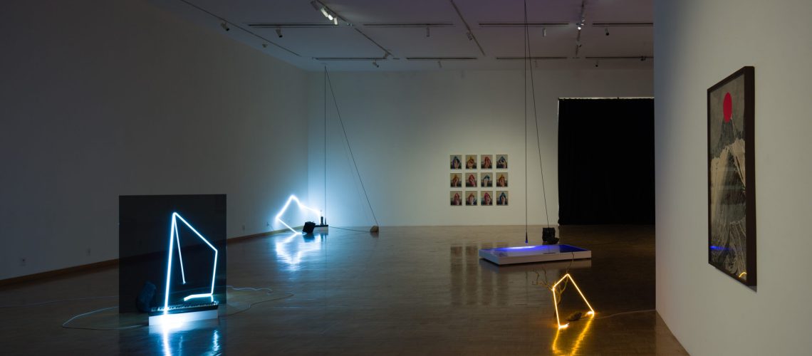 A spacious art gallery room with wooden flooring showcasing an eclectic mix of modern artworks, including neon light installations, framed pieces on the walls, and a projected image.