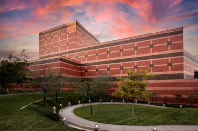 A modern brick building with a geometric design is surrounded by a neatly landscaped lawn and trees. A curved pathway leads through the greenery. The sky is vibrant with hues of pink and orange at sunset.