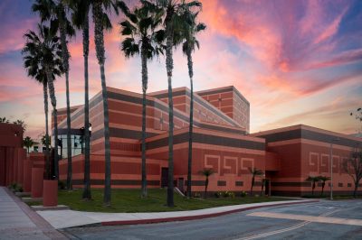 A modern building with geometric patterns and a red brick exterior, surrounded by tall palm trees. The sky is colorful with shades of pink, orange, and blue, suggesting a sunset or sunrise. A deserted street curves around the building.