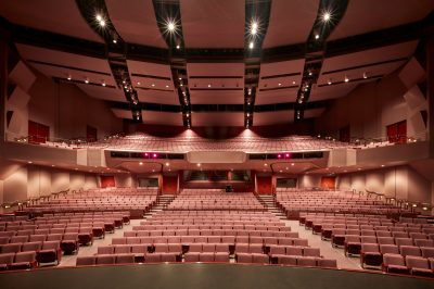 A large, empty theater with rows of red seats, a spacious stage in the foreground, and dim overhead lighting. The auditorium features two balconies and a central aisle, creating a grand, symmetrical view from the stage.