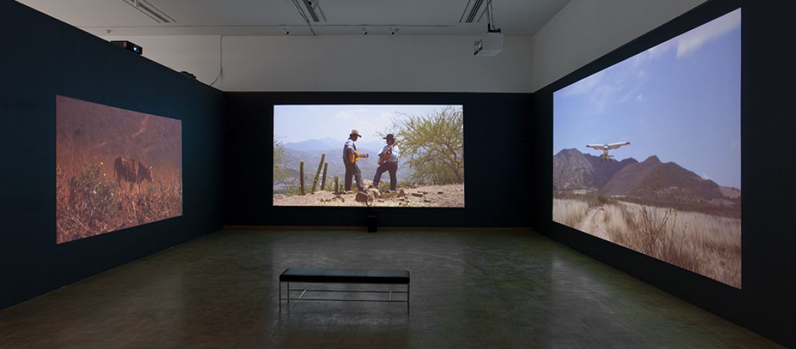 An immersive multi-screen video installation in a dark gallery space, featuring natural landscapes and human interaction with the environment, inviting viewers to a contemplative experience.