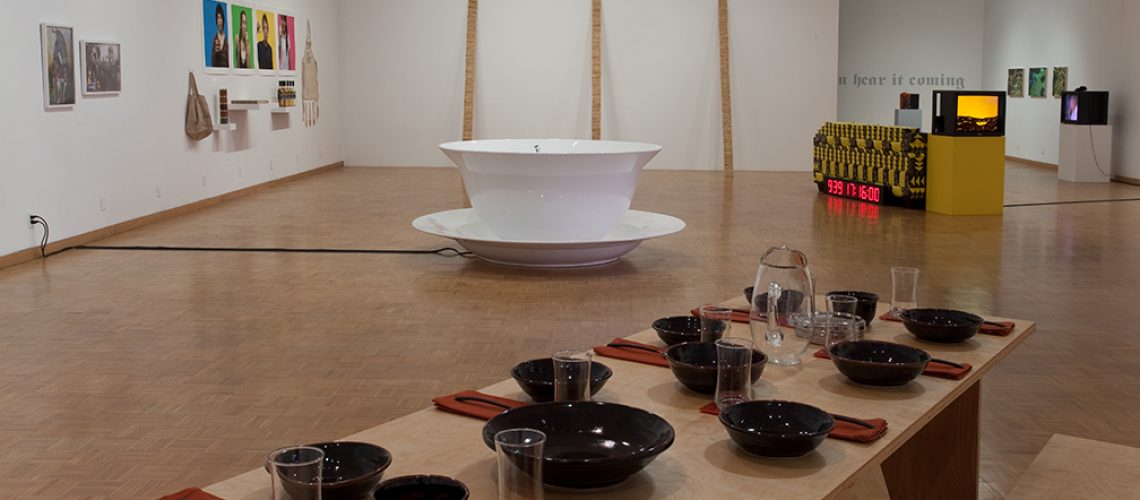 An art gallery displaying various installations, featuring a large white basin in the center surrounded by wooden poles, a table set with black bowls and glasses, and colorful wall pieces in the background.