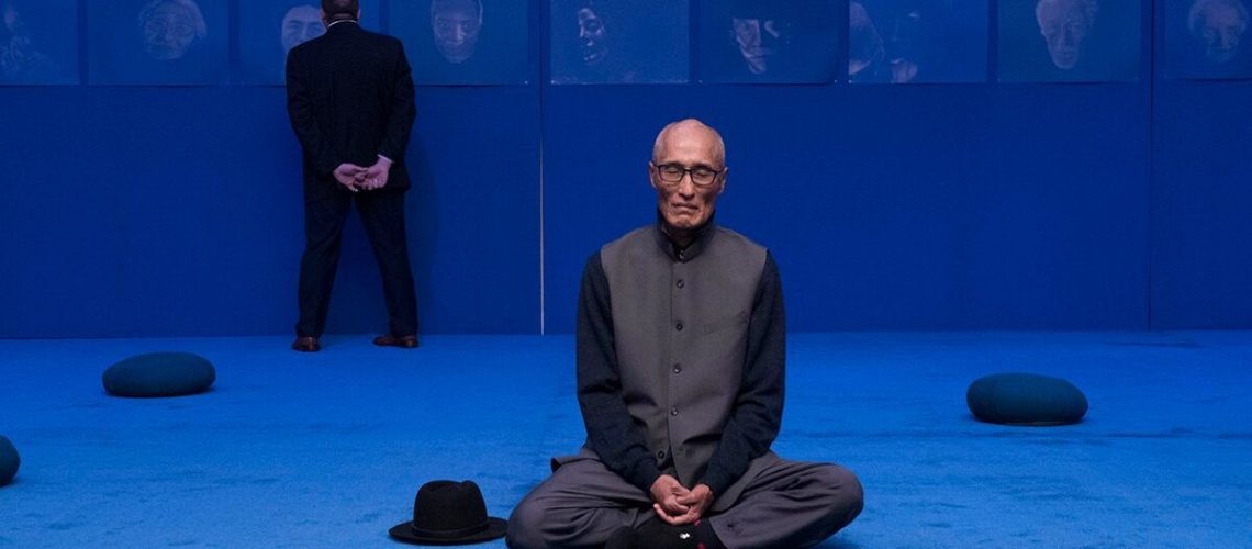 Ying Ming Tu in a dark tunic sitting cross-legged on a vibrant blue carpeted floor with a hat placed beside him, while another person stands in the background looking at framed portraits on the wall.