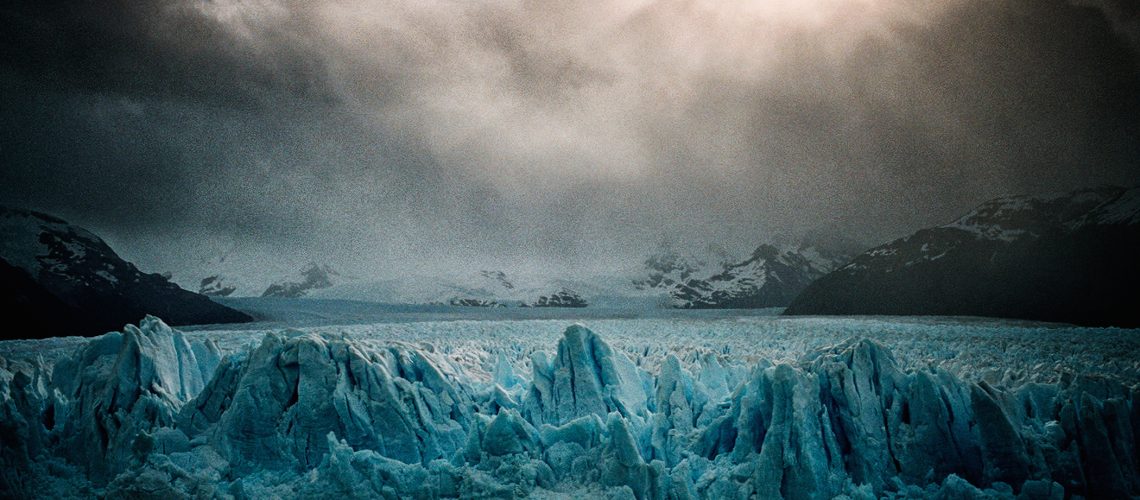 A dramatic scene of a sprawling glacier under a moody sky pierced by the glow of the sun, captured in the unique style of Bruce Yonemoto.