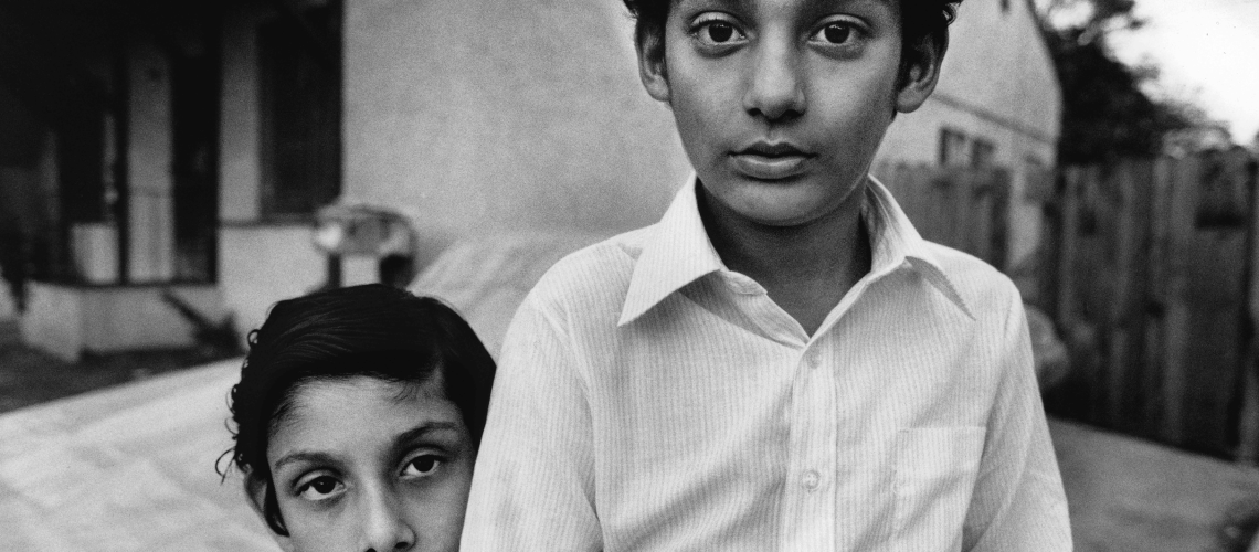 Two boys with earnest expressions, one resting his head on the other's shoulder, share a moment of camaraderie in a stark, monochromatic setting.