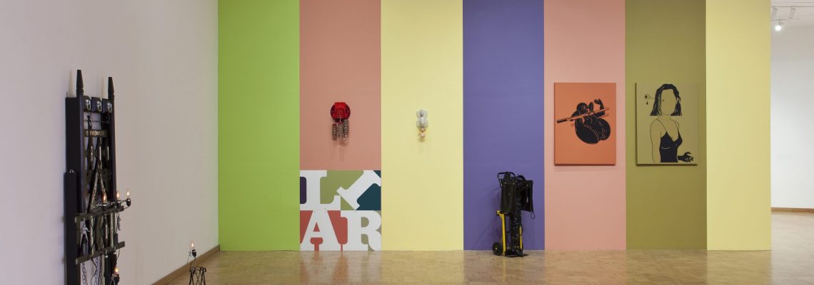 A modern art gallery with lively colored walls featuring a rainbow of vertical panels – green, yellow, blue, purple, and pink – adorned with eclectic artwork, sculptures, and some photography equipment on the wooden floor.