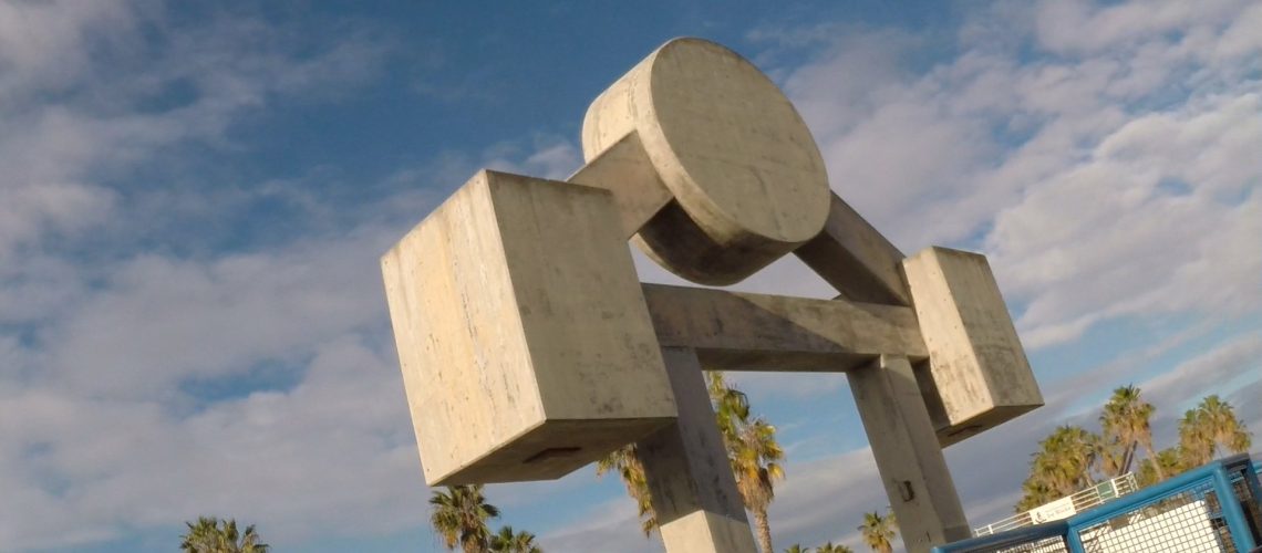 A low-angle view of a modern concrete sculpture with geometric shapes, set against a backdrop of a clear blue sky with fluffy clouds and framed by tall palm trees.