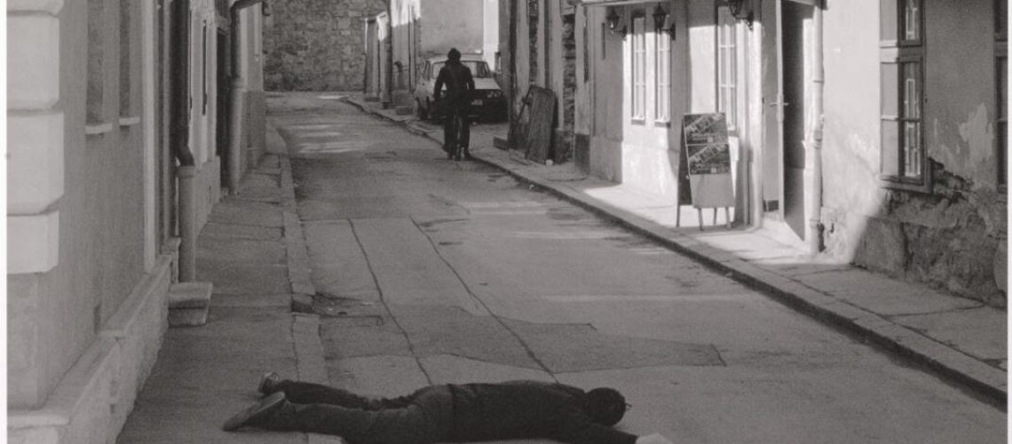 A person lying face-down on an empty street in a seemingly peaceful town, while another figure walks away in the distance.