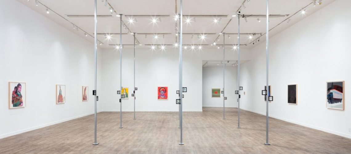 A contemporary art gallery interior displaying various paintings on white walls, with a unique installation of hanging frames in the center, under bright ceiling lights.