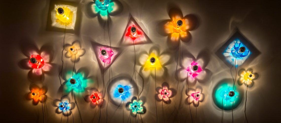 Colorful flower-shaped wall lights in various colors creating a vibrant and whimsical display against a dark background with soft shadows.
